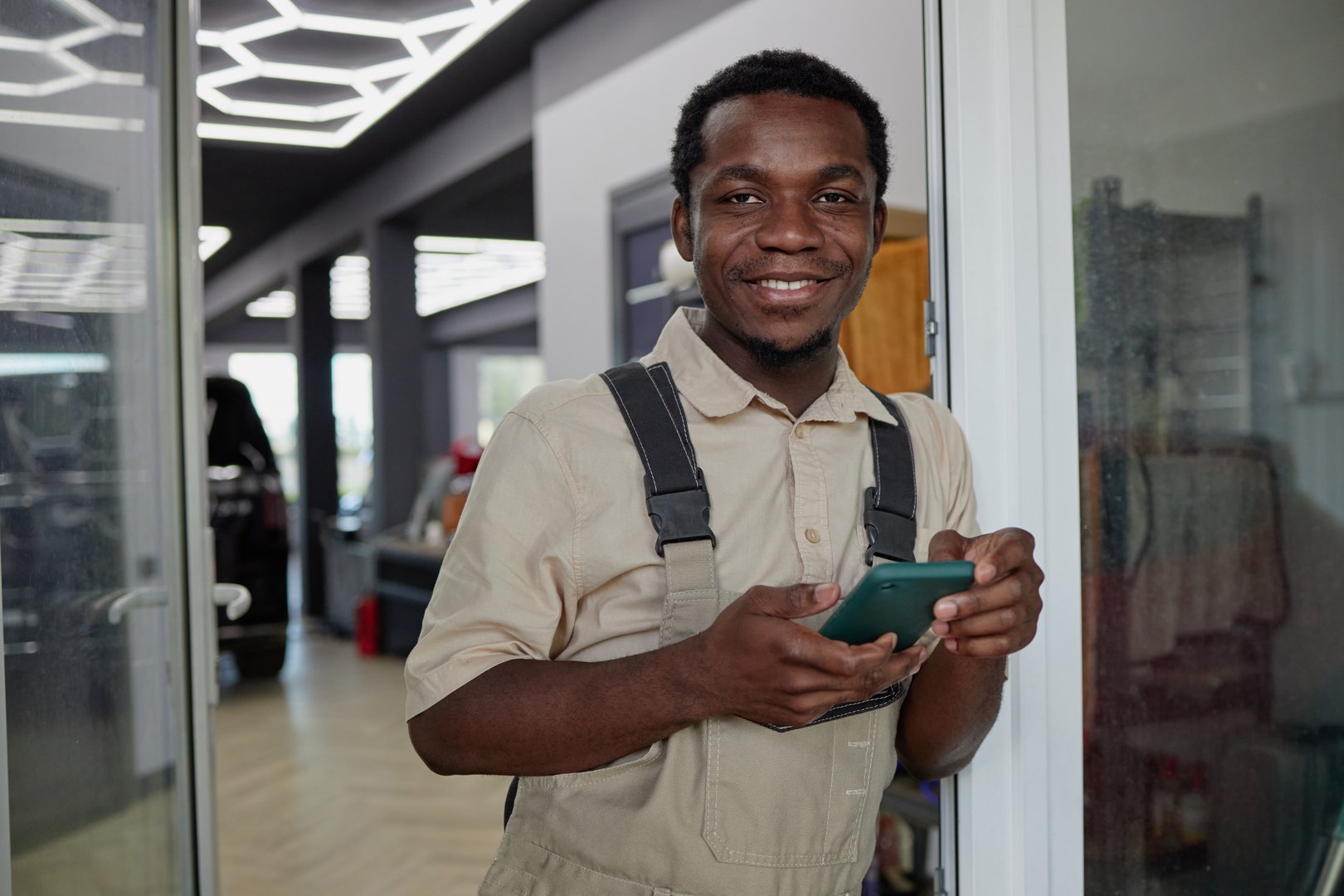 Portrait of young adult Black man smiling while holding smartphone, standing in modern indoor setting near glass door, wearing work overalls, looking directly at camera