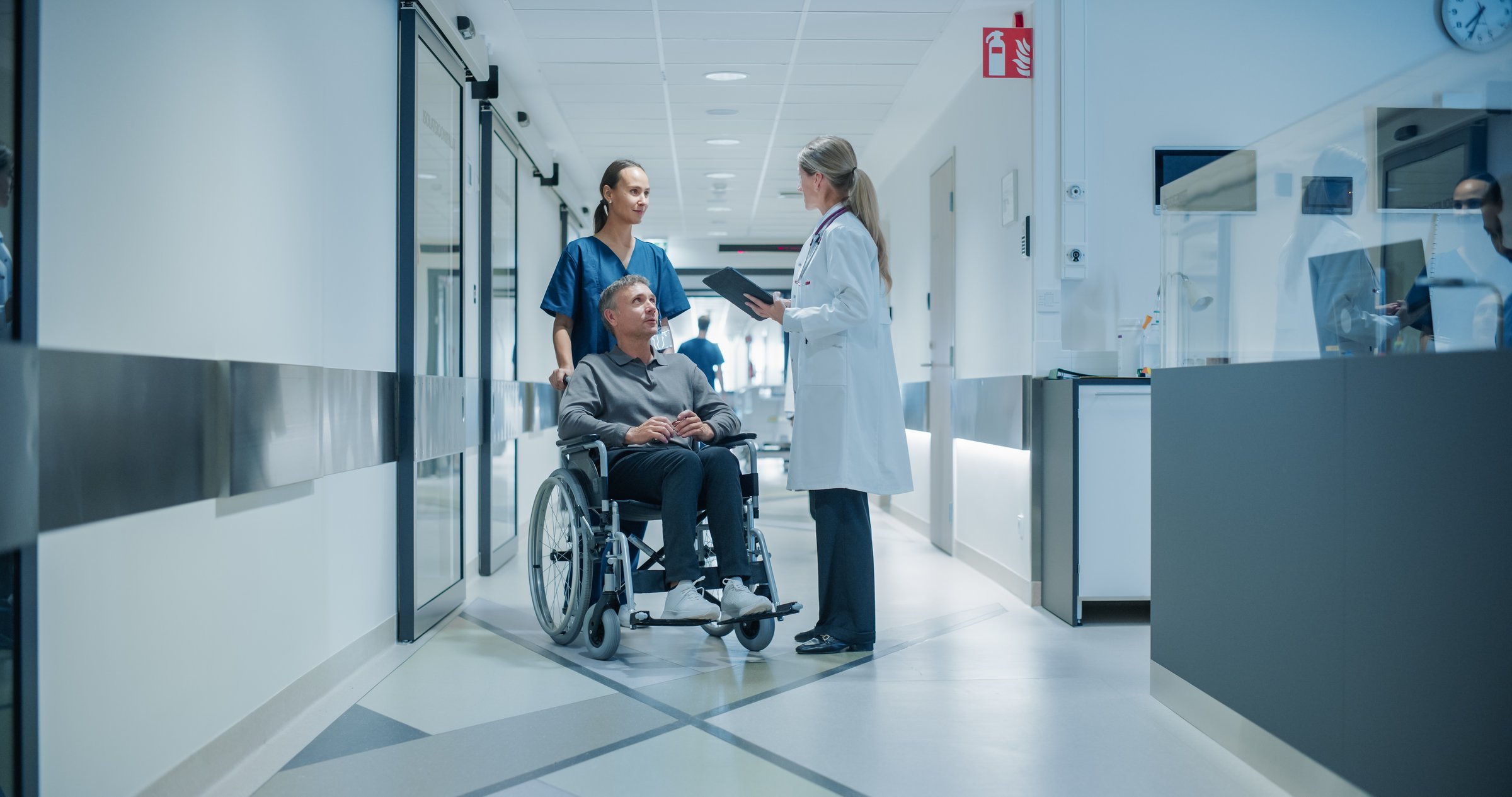 Female Doctor Approaching a Male Patient in a Wheelchair in a Bright Hospital Corridor Next to a Registration Desk, Having in a Friendly Conversation while Reviewing Notes on a Tablet Computer
