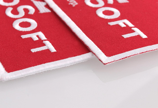Two red fabric patches with embroidered white text and white borders, overlapping on a smooth, reflective surface.