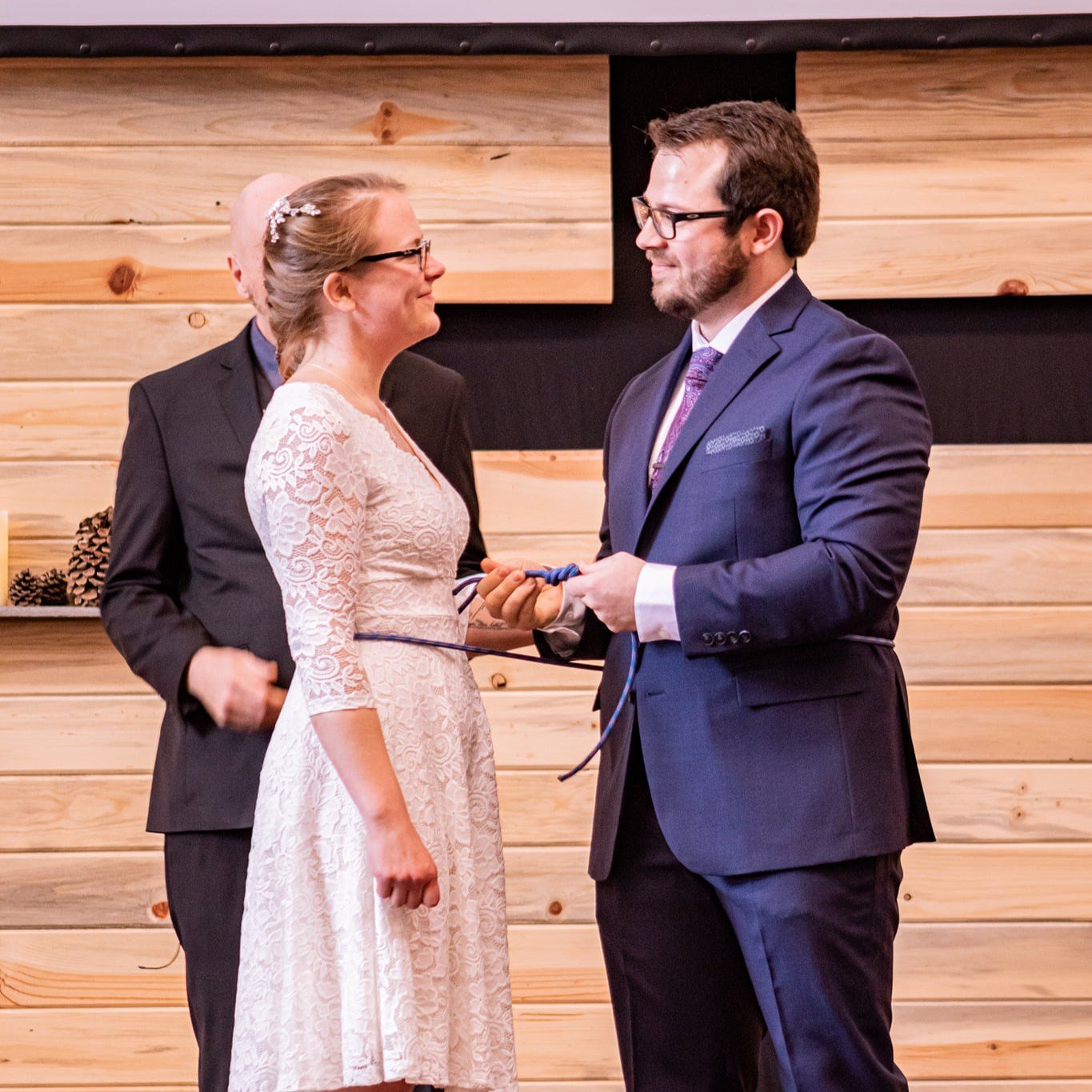 A man and woman holding hands at a ceremony, each wearing a suit and white dress, with a wooden backdrop behind them.