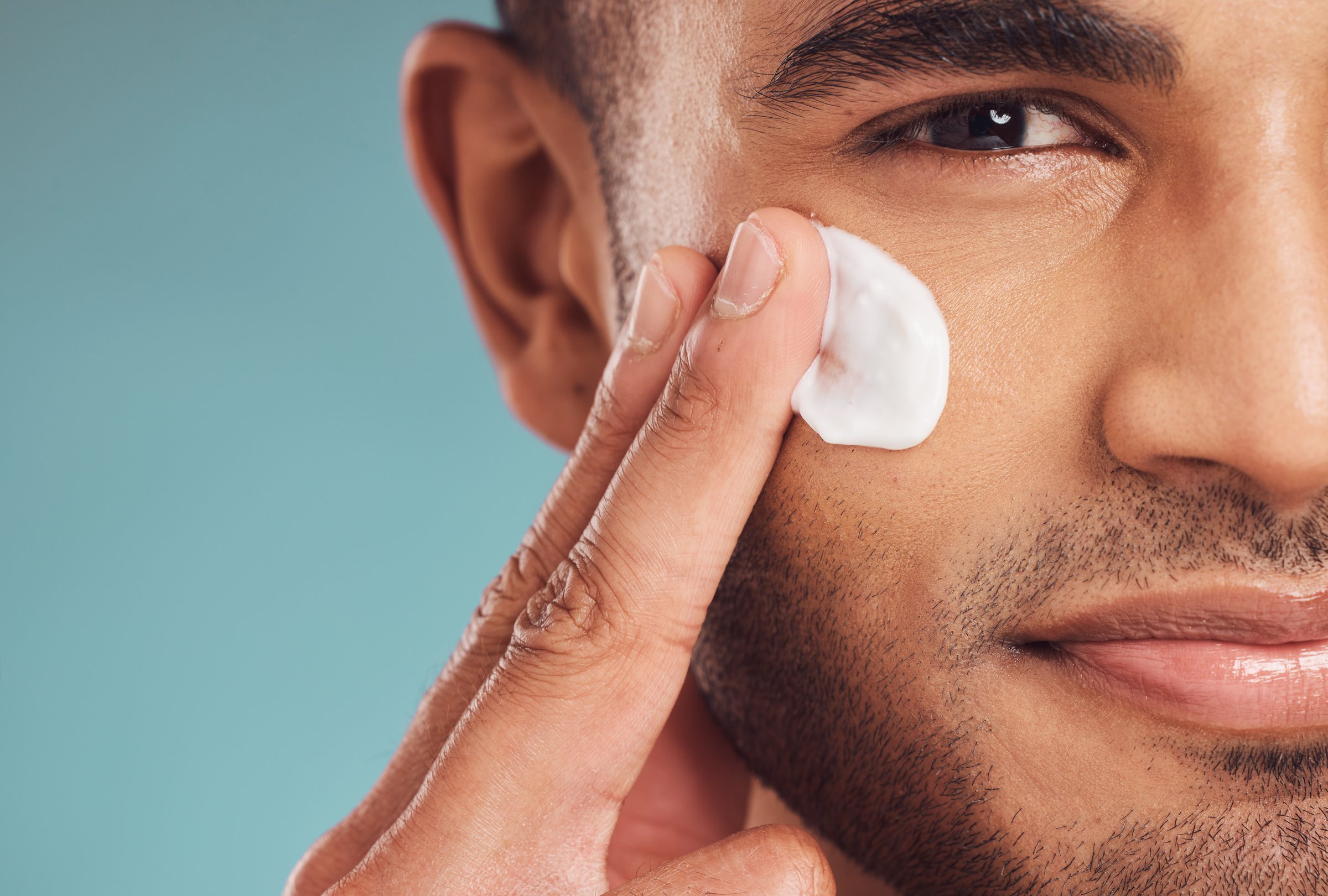 Closeup of one young indian man applying moisturiser lotion to his face while grooming against a blue studio background. Handsome guy using sunscreen with spf for uv protection. Rubbing facial cream on cheek for healthy complexion and clear skin