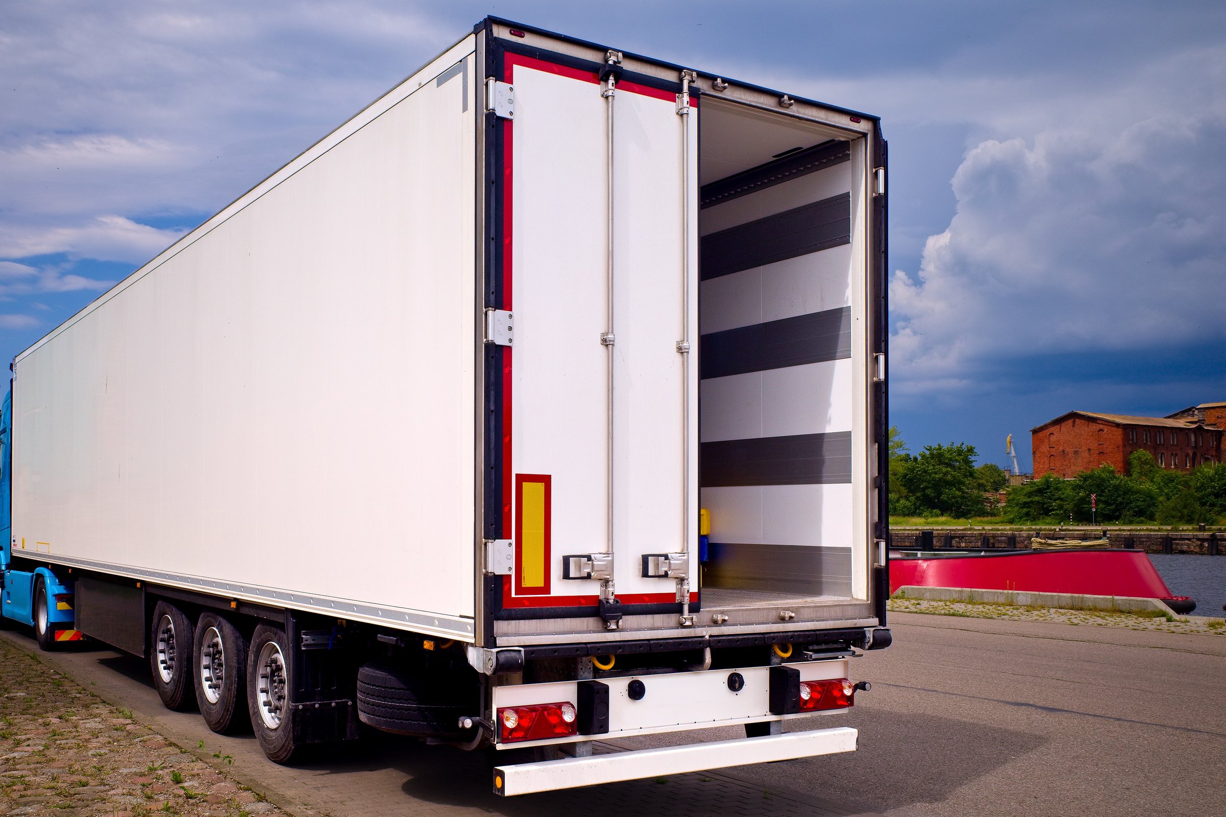 Refrigerated truck trailer with open rear doors parked on an industrial street near a warehouse and river port, ready for cargo loading.