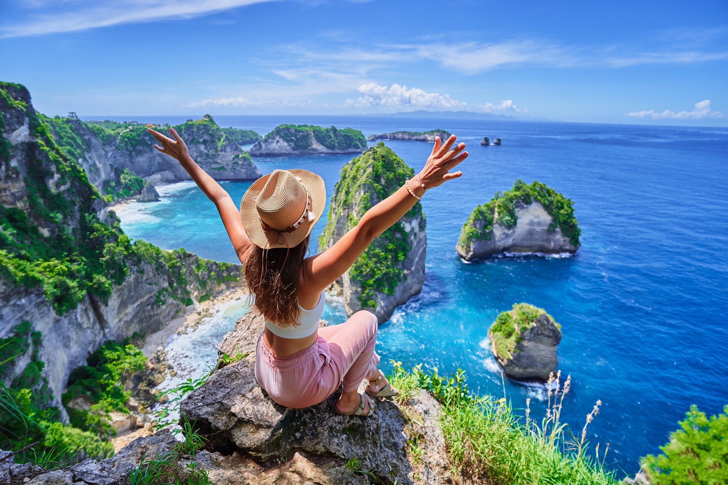Woman traveler with open arms sitting on rock cliff and enjoying of scenic landscape of Diamond beach during vacation to Nusa Penida Island in Indonesia