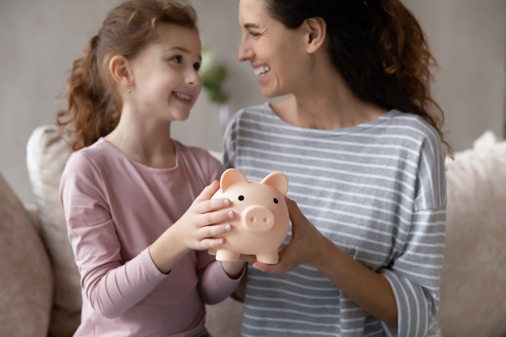 Close up happy mother with smiling little daughter holding pink piggy bank sitting on couch at home, caring mom and adorable girl child saving money for future, family insurance and investment