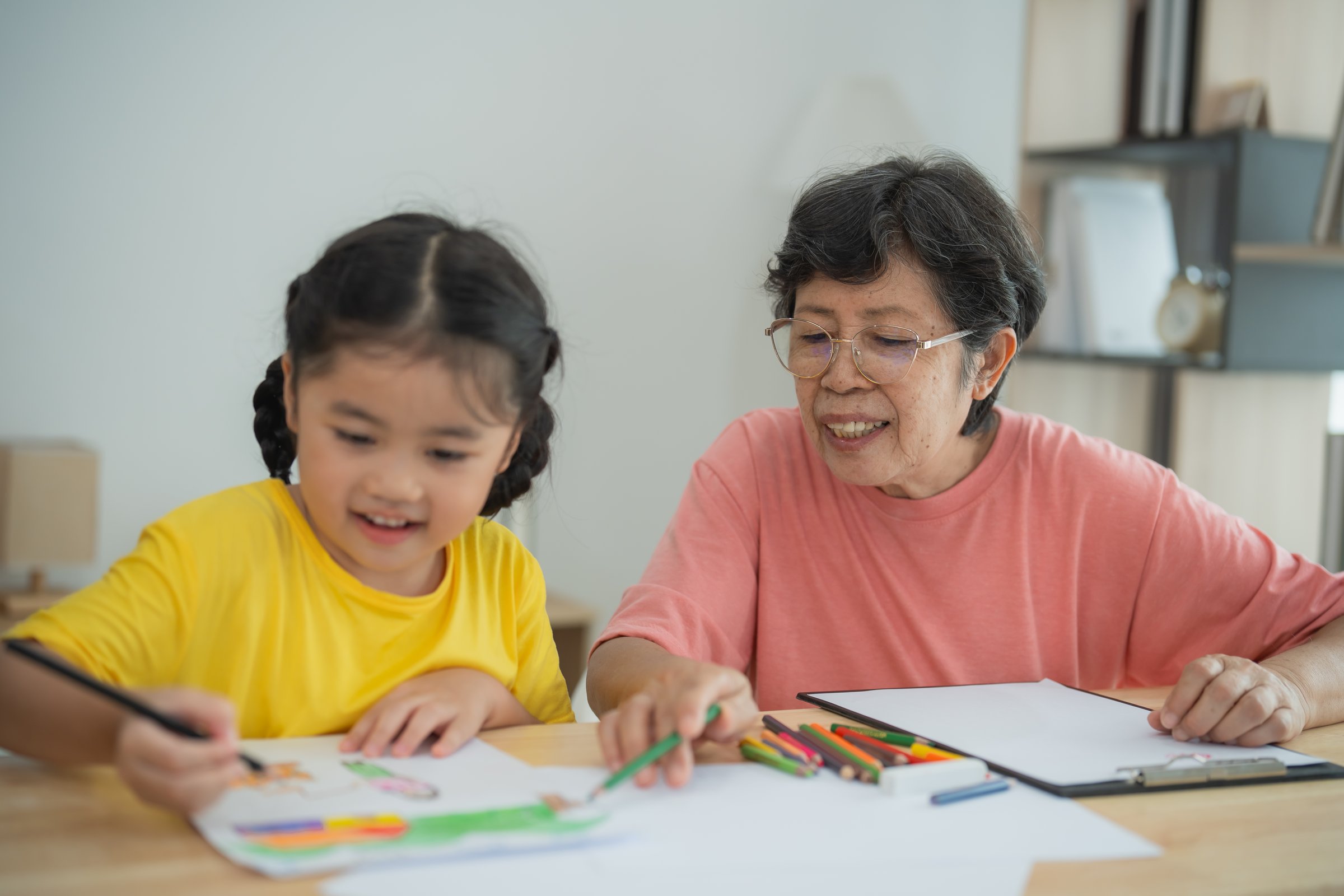 Asian cheerful grandmother and her granddaughter share a delightful moment while creating colorful artwork together, showcasing the beauty of family bonds and creativity in a bright indoor space.
