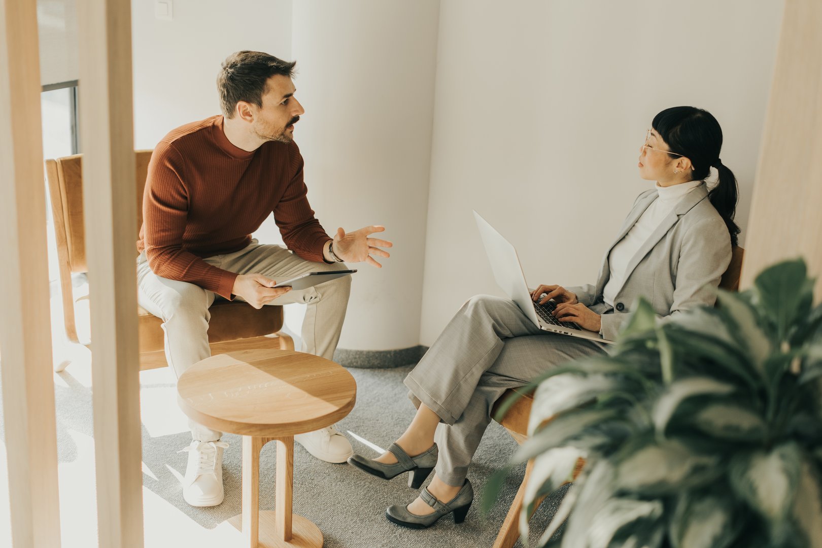 Two professionals engage in a thoughtful discussion in a stylish office setting, exchanging ideas while seated comfortably in contemporary chairs.