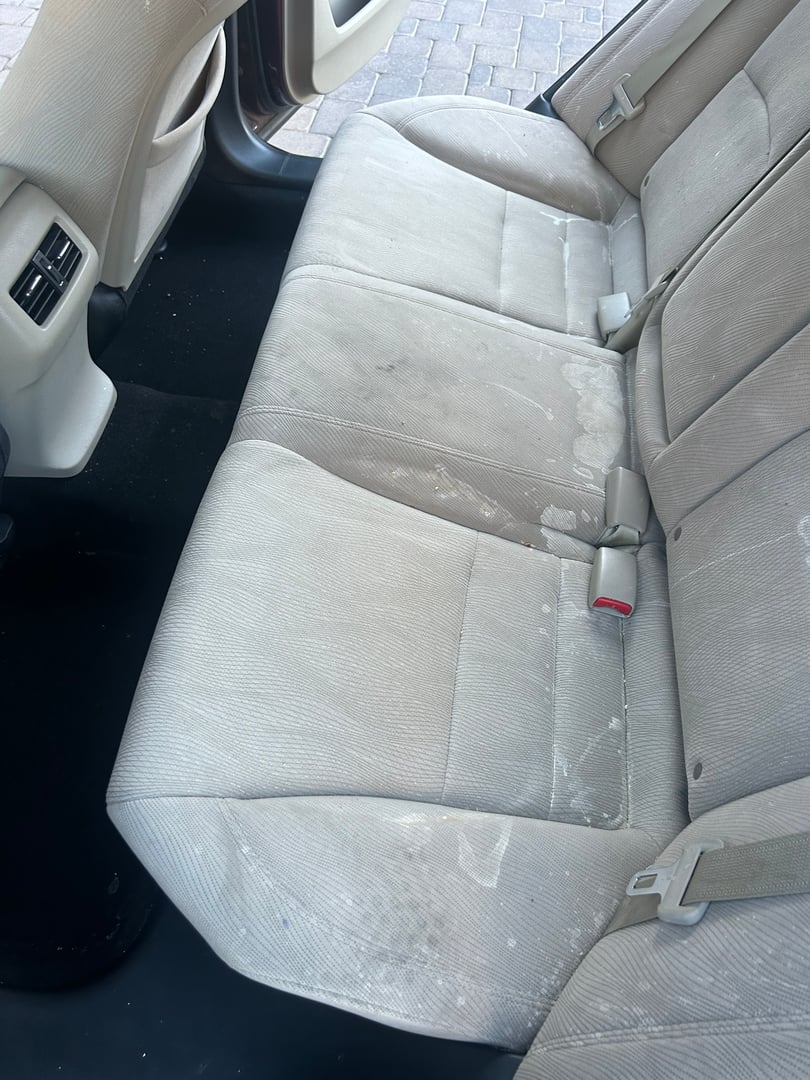 Car's back seat with beige upholstery showing visible stains and some leftover debris, suggesting recent use or spill.