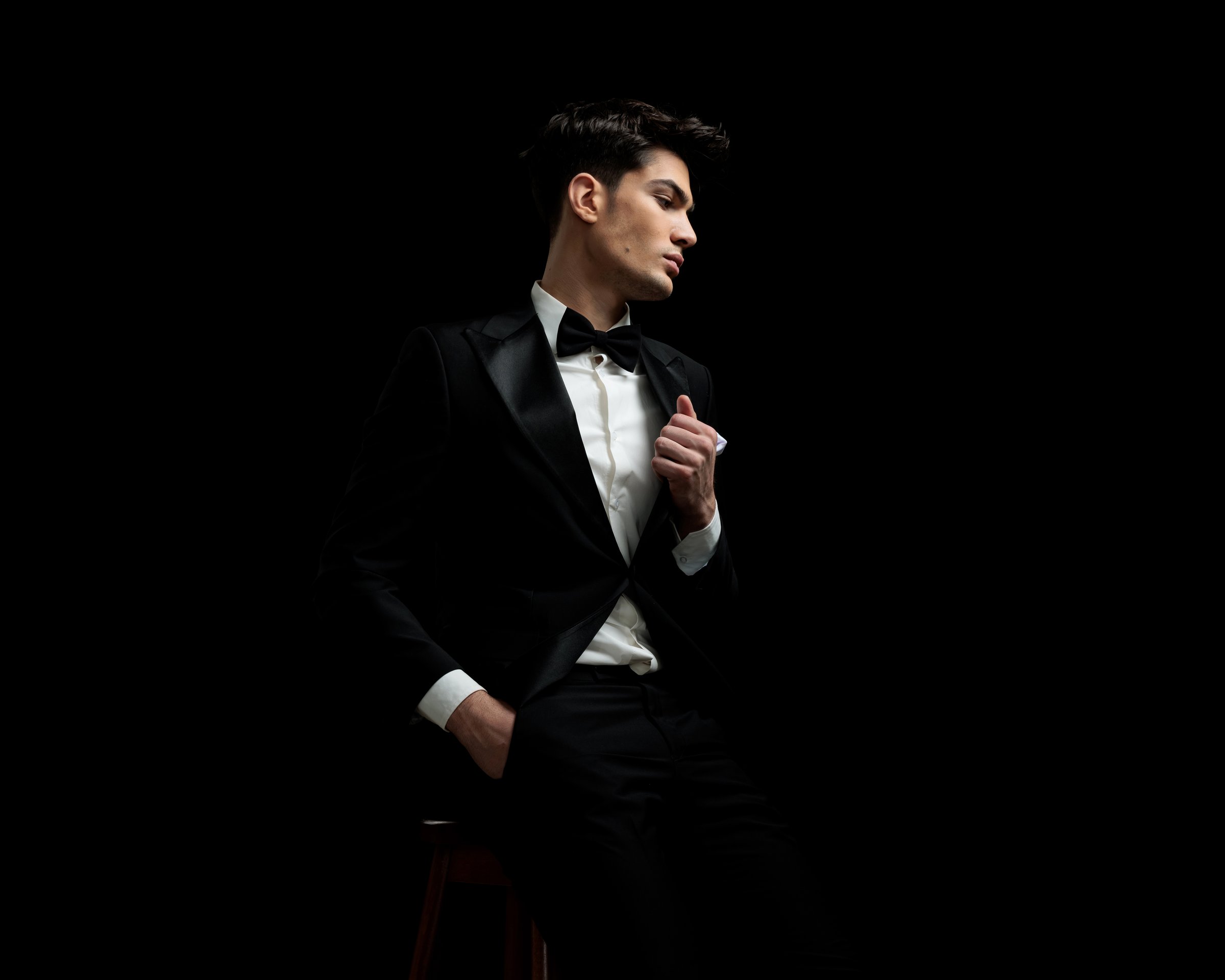 sexy fashion man holding pocket and fixing black suit collar while looking down to side on black background