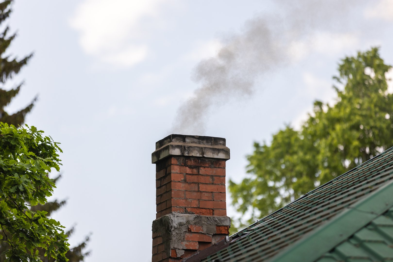 A brick chimney releases smoke over a green-tiled roof in a calm suburban scene. Trees frame the background as a gentle plume rises, suggesting warmth, home, and outdoor weather