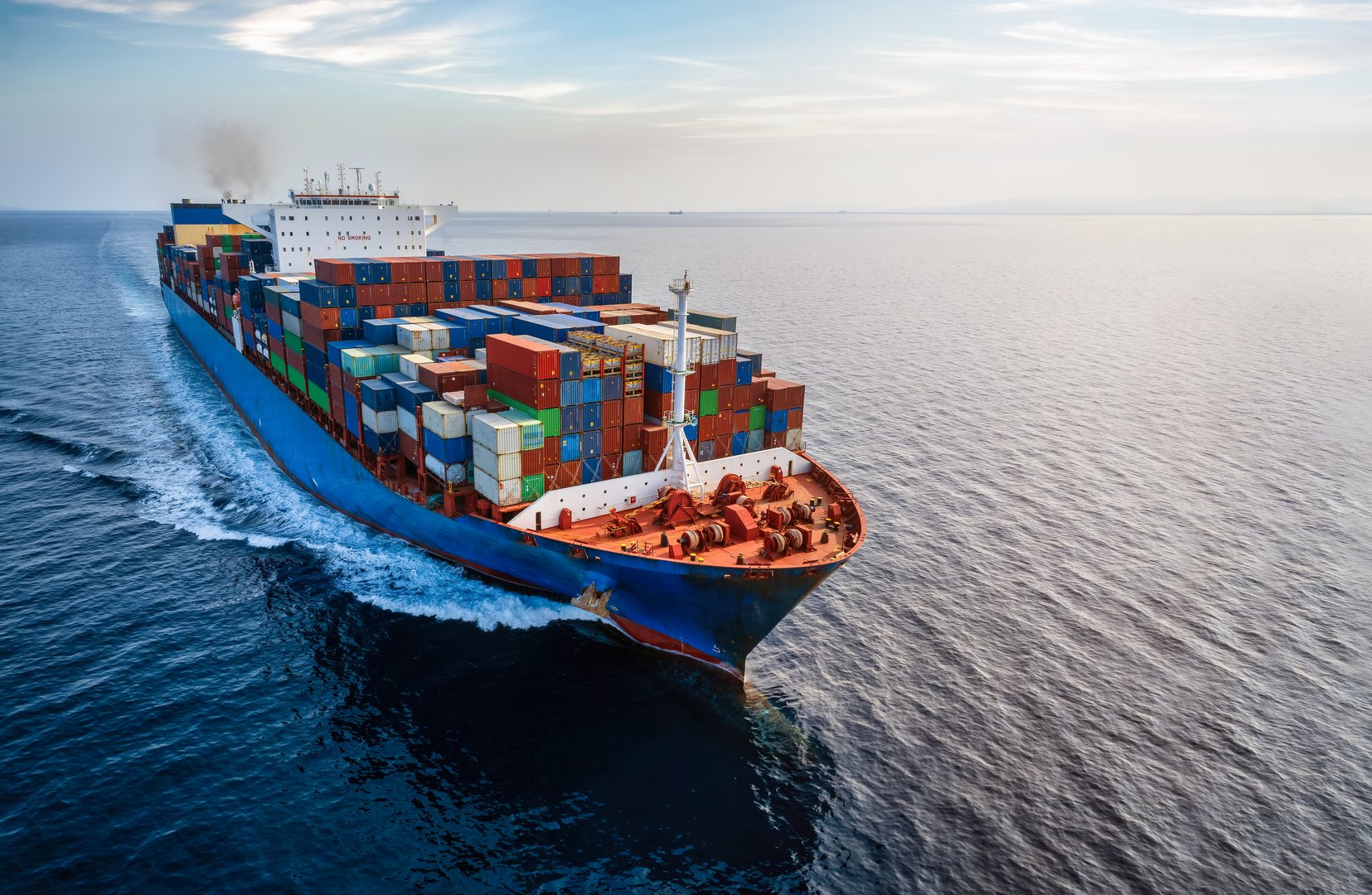 Aerial front view of a large container cargo ship traveling with speed over calm ocean and sunset light