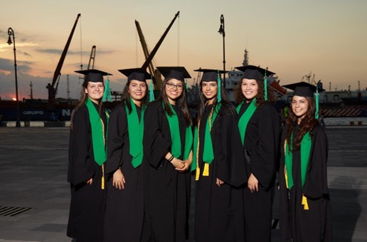 Six graduates in caps and gowns with green stoles pose for a photo outdoors, with cranes and a sunset in the background.
