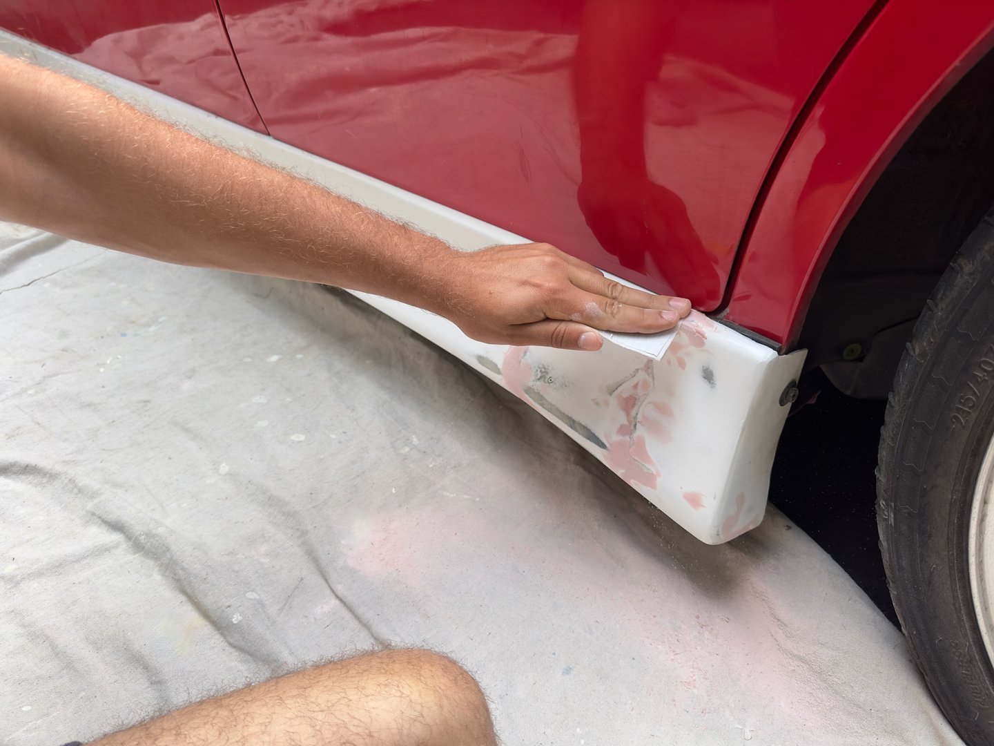 Close up of a male hand holding a small piece of sandpaper to smooth the pink bog patches on the white skirt of a red car body