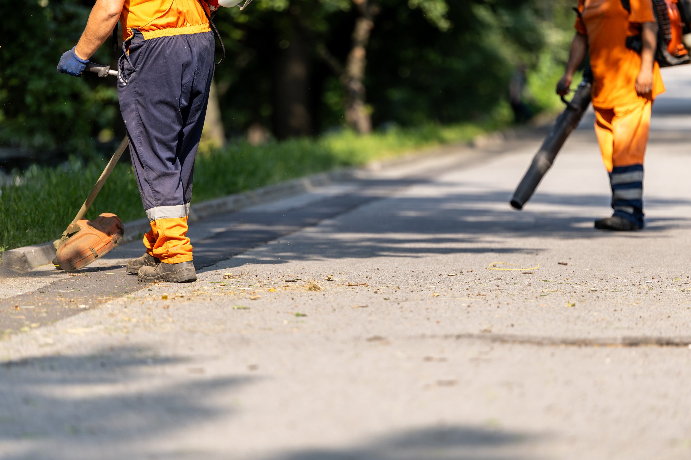 Two workers in orange uniforms are performing landscape maintenance in a park. One is using a trimmer while the other operates a leaf blower, ensuring the area remains clean and tidy.