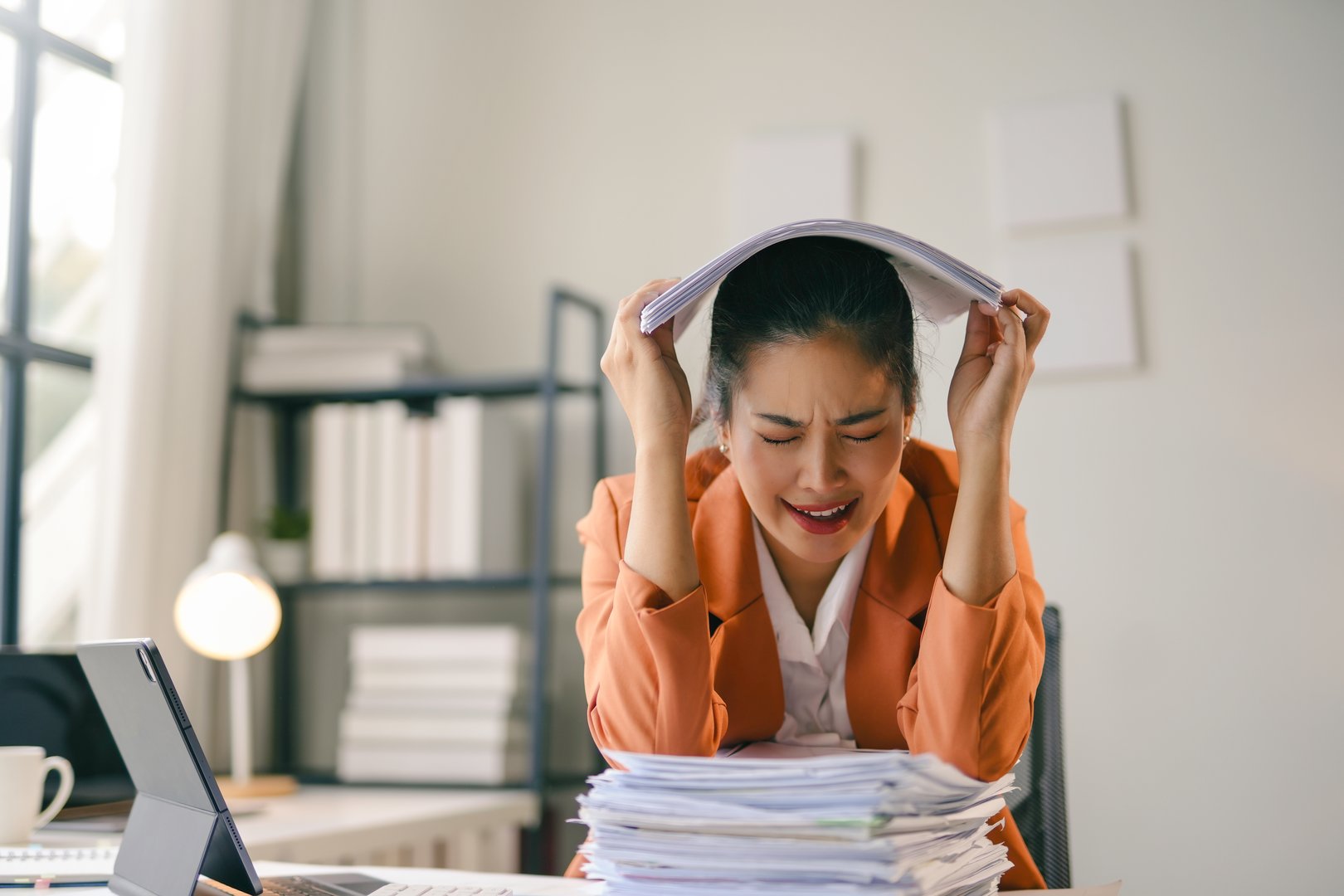 Stressed asian businesswoman holding documents over her head while sitting at her desk with a large pile of paperwork in front of her, feeling overwhelmed and frustrated