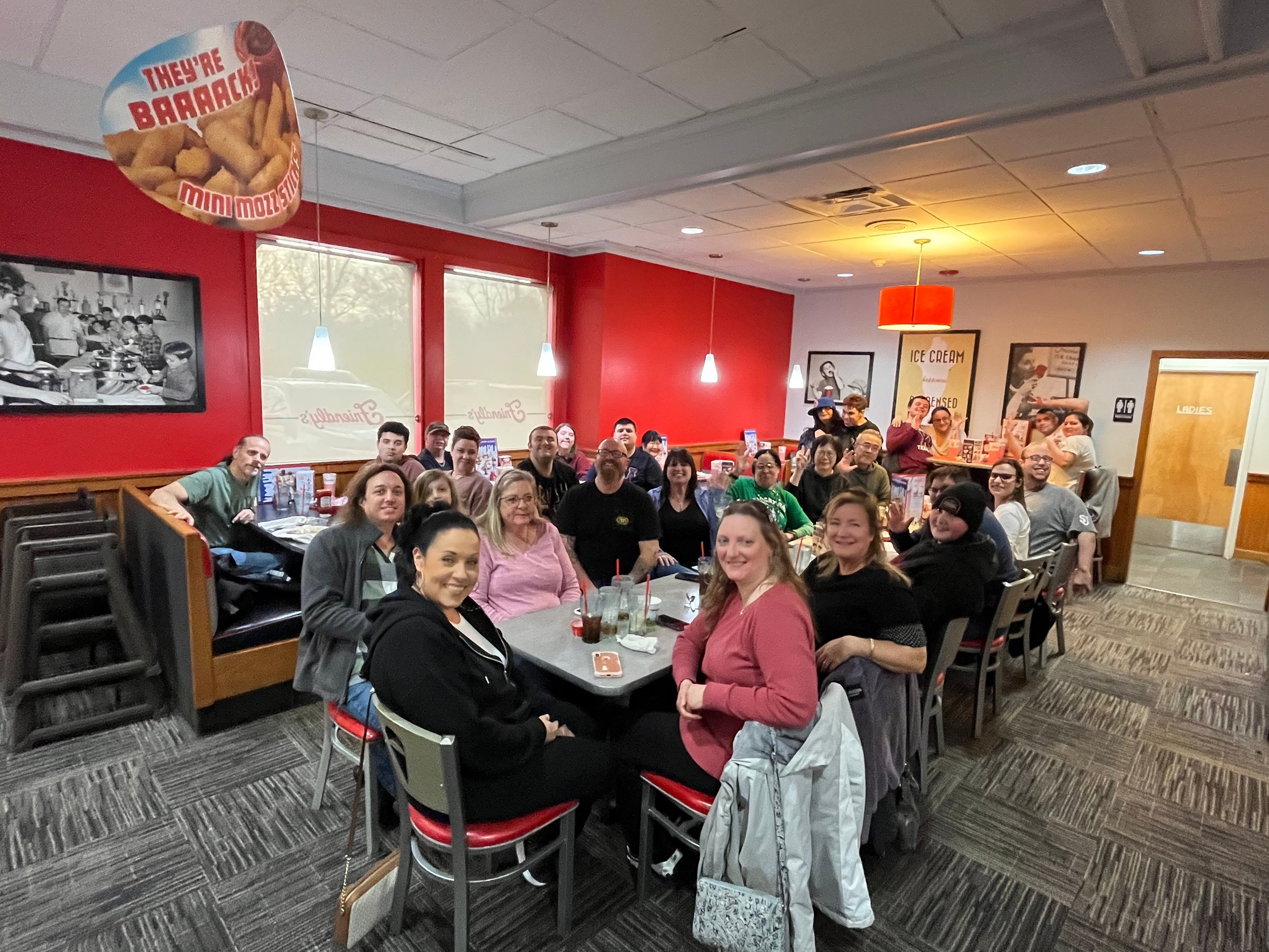 A large group of people seated around tables in a diner-style restaurant, smiling at the camera. Red walls and retro decor visible.