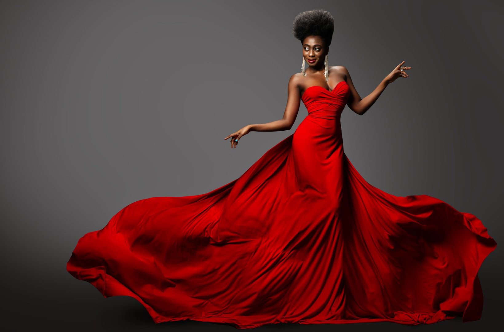 Fashion Model in Red Long waving Luxury Dress. Dark Skinned Beauty Woman with Afro Black Hairstyle dancing over Gray Background. Happy African Sexy Girl in Silk Evening Gown with flying Fabric