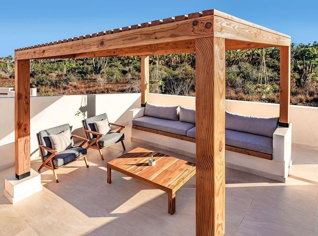 Outdoor patio with a wooden pergola, cushioned seating, two chairs, and a coffee table on a tiled floor.