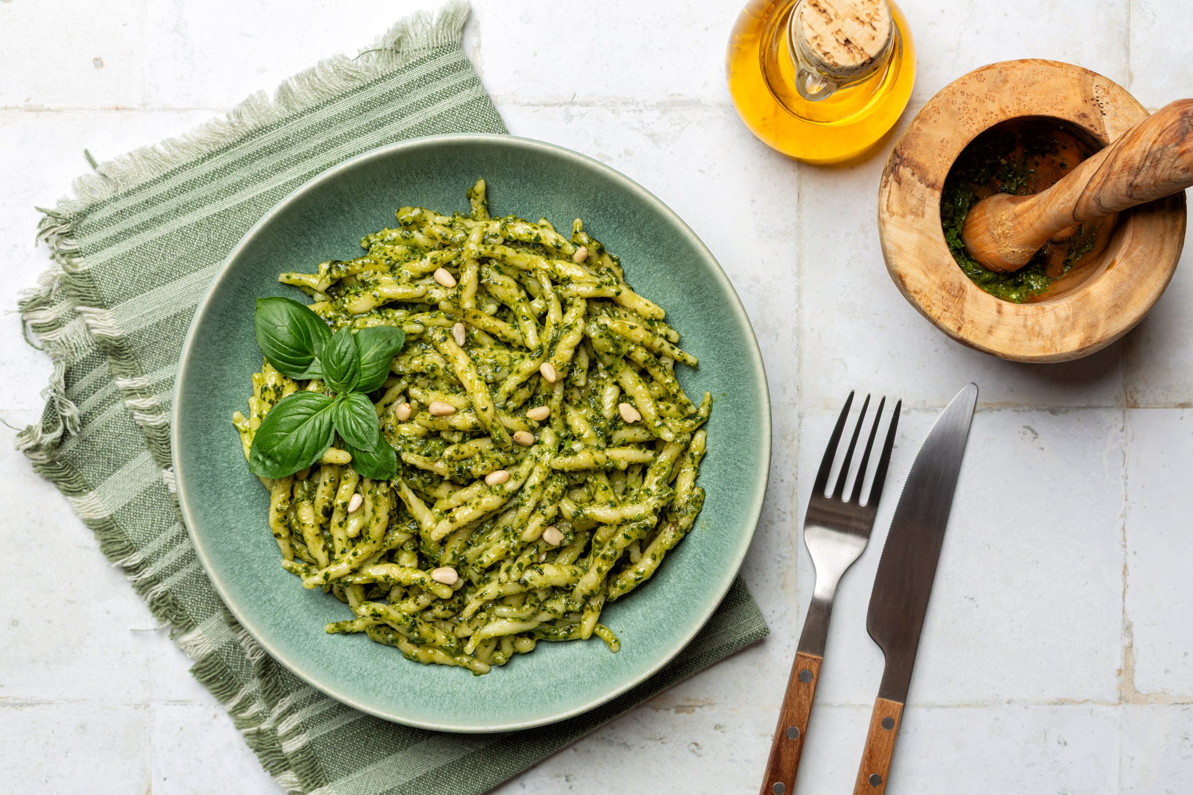 Top view of plate with Trofie al pesto,  pasta with homemade pesto genovese, made with basil leaves, olive oil, parmesan, pecorino, pine nuts, and garlic. White table surface, greeb napkin.