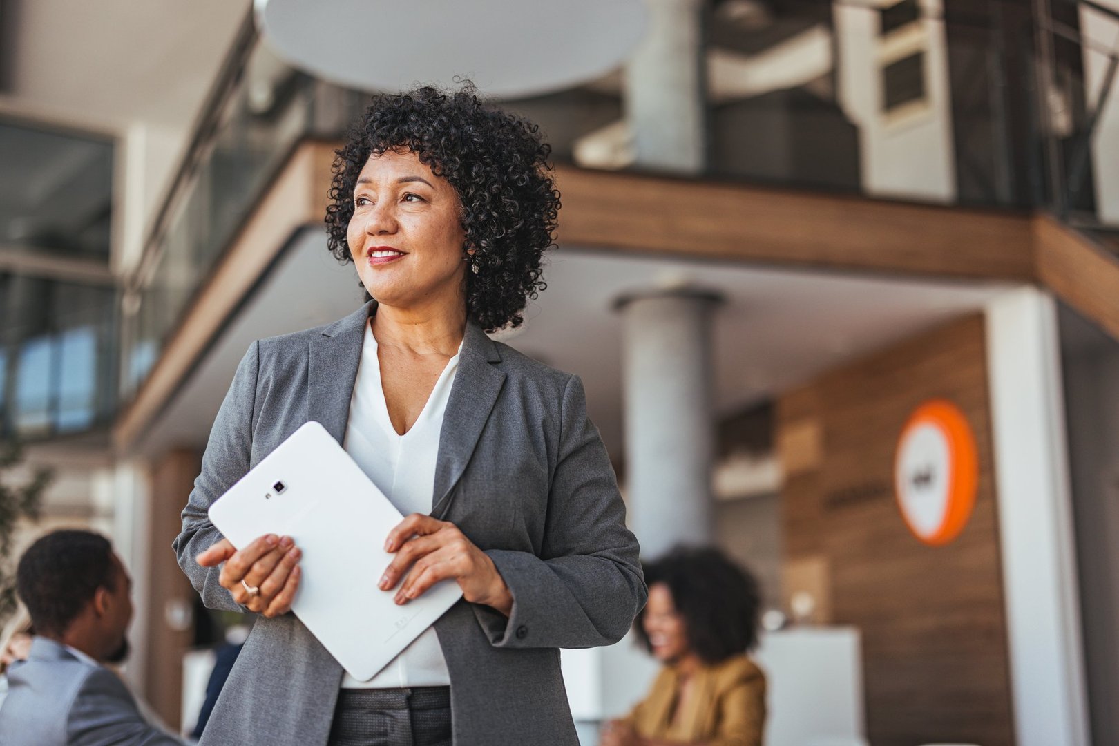A confident businesswoman stands holding a tablet in a modern office. The setting conveys professionalism and success. Background includes colleagues, reflecting a collaborative and dynamic work environment.