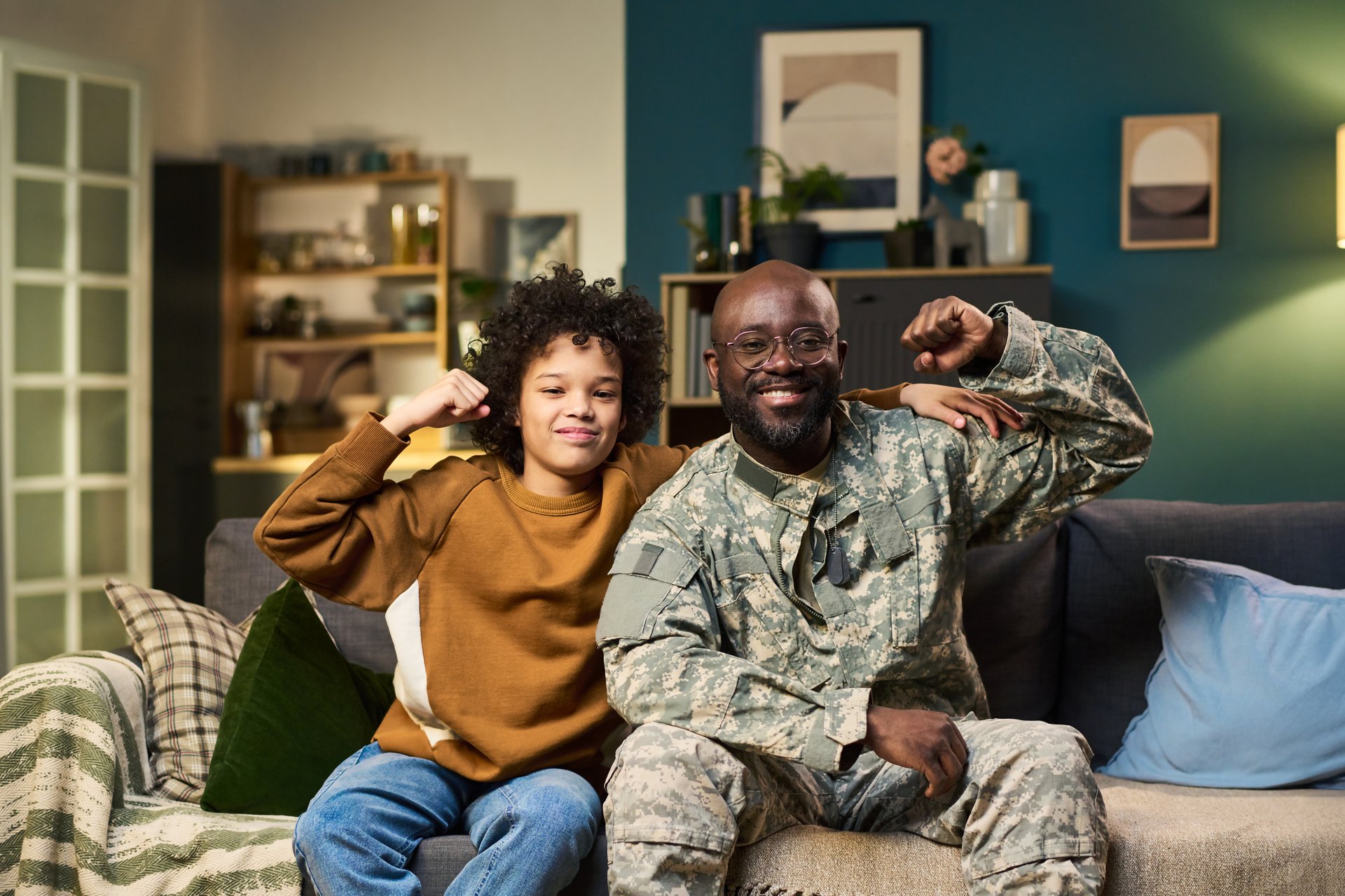 Black middle aged man in military uniform sitting on couch with Black child flexing arms and smiling together at home, both looking at camera, showing family bond and pride