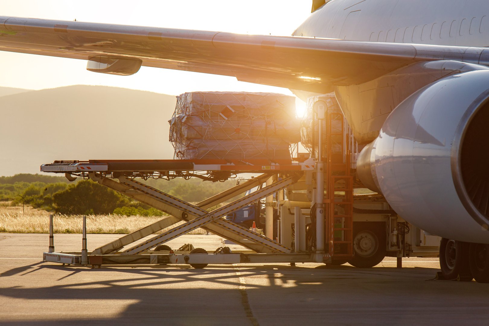 Workers load cargo onto a commercial airplane as the sun sets behind the mountains.