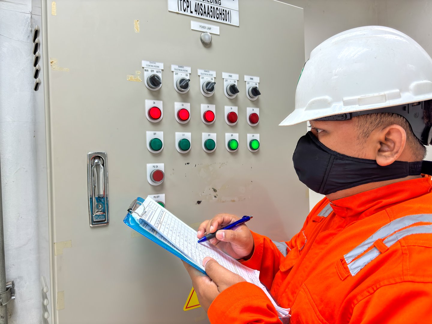 A skilled industrial worker wearing safety gear meticulously inspects a control panel making notes on a clipboard ensuring adherence to operational standards and regulations in an industrial setting.