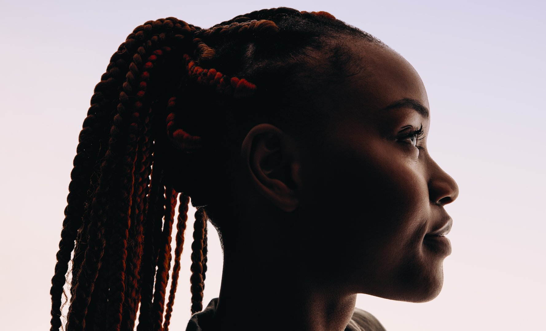Young African woman with braided hair styled in a ponytail. She is seen in a closeup headshot, looking away with a side view. The white background emphasizes her stylish hairstyle and exudes her strong sense of confidence.