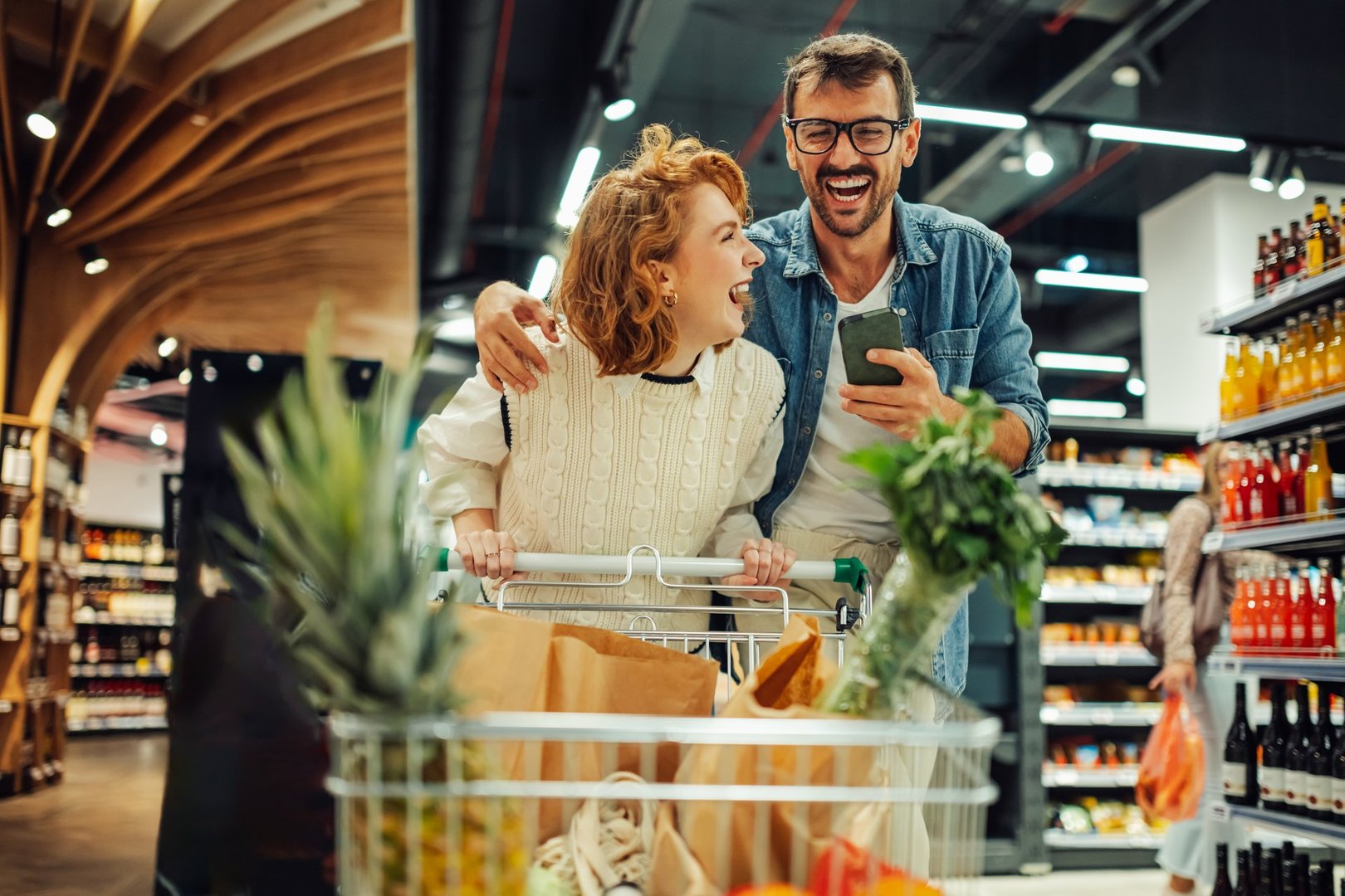 Young couple laughs while using a smartphone in a supermarket, pushing a cart filled with healthy groceries