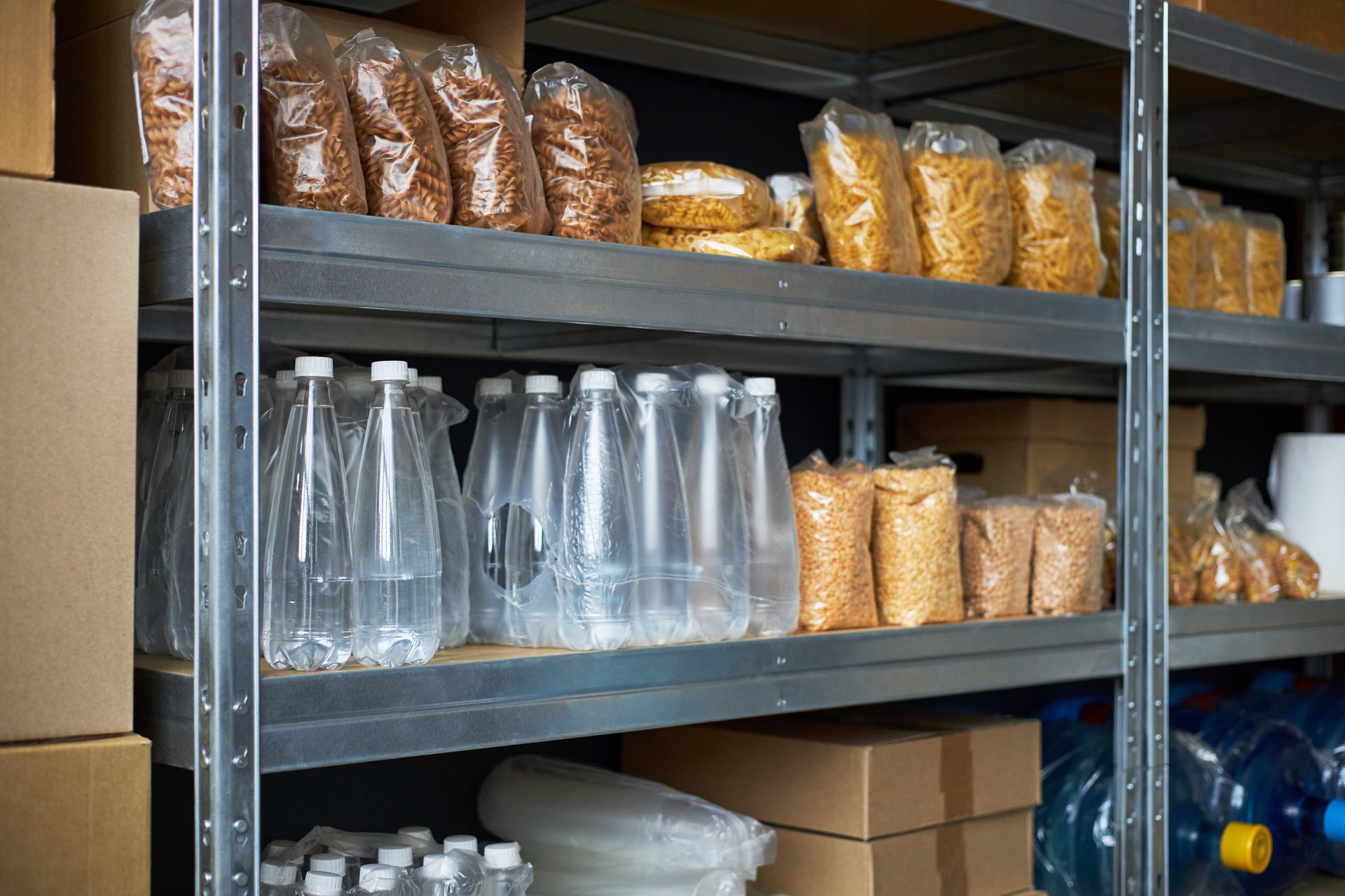 Shelves filled with packaged food and bottled water in a storage area. Supplies neatly organized on metal racks with various items including pasta and snacks