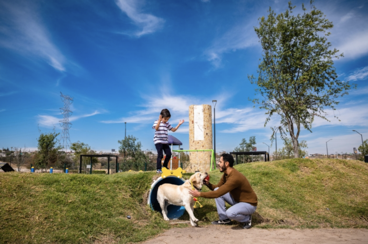 Man with dog greets girl coming out of a colorful play tube in a park, under a clear blue sky.