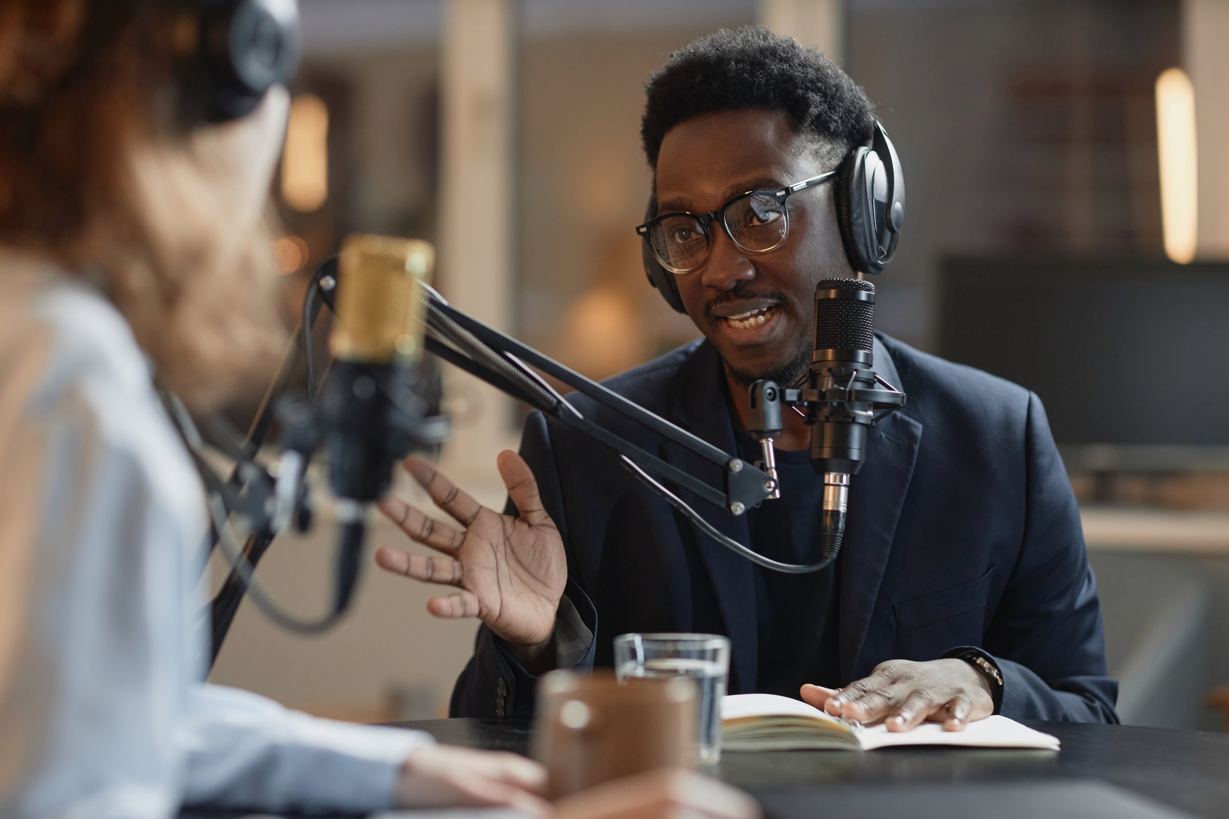 Medium shot of young Black man in eyeglasses and suit answering questions of unrecognizable interviewer during podcast recording