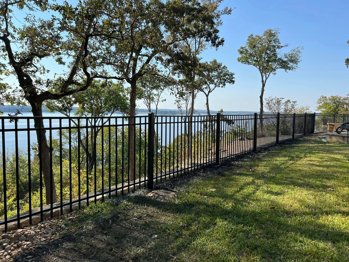 A black metal fence runs alongside a grassy area with trees in the background and a view of a distant lake under a clear blue sky.