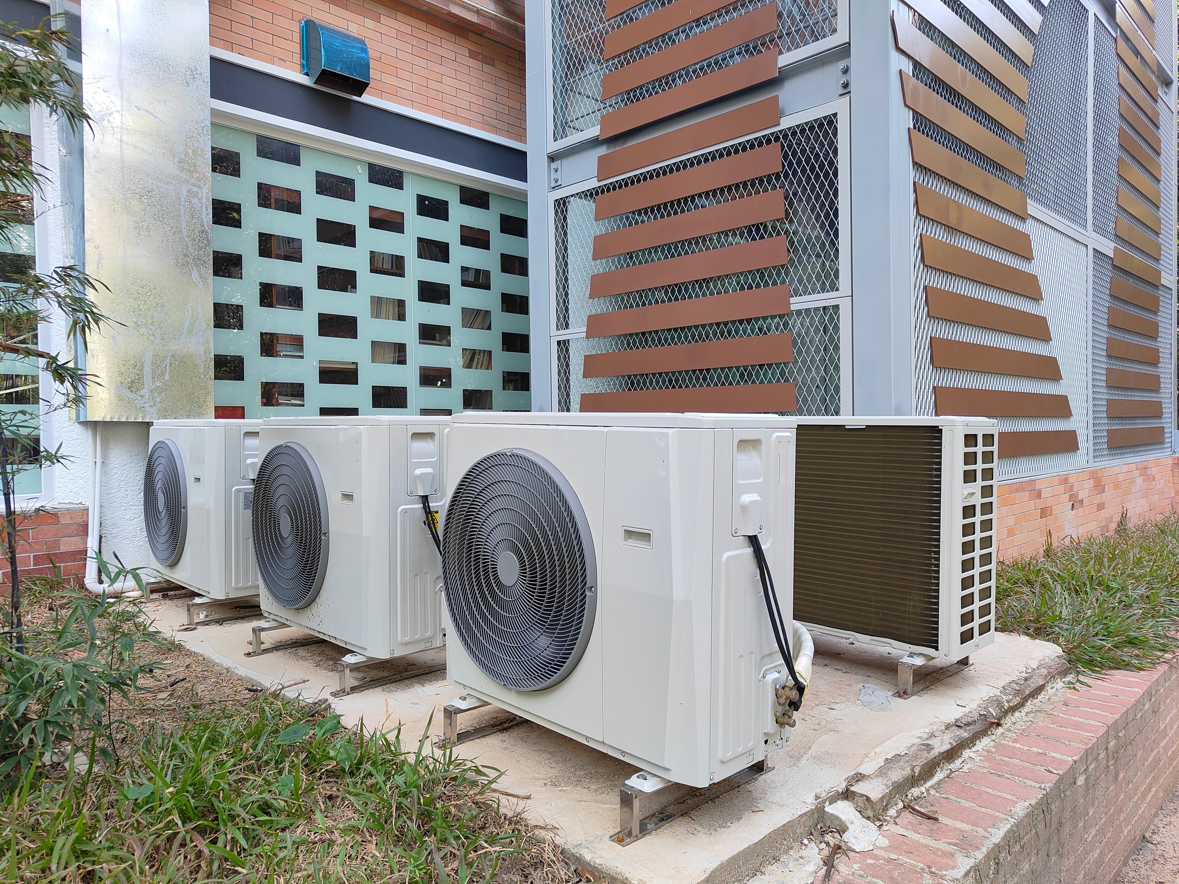 Three air conditioners are mounted on a wall. The air conditioners are white and black. The wall is made of bricks