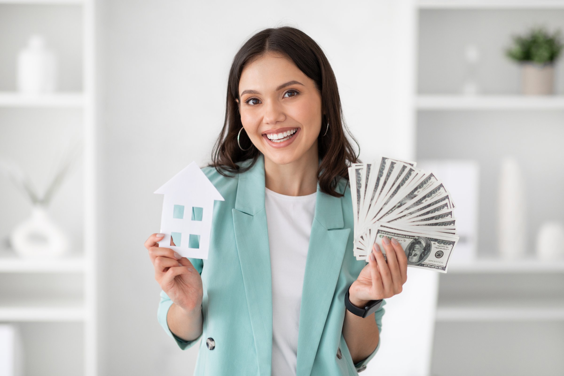 Smiling millennial caucasian lady manager in suit hold a lot of dollars cash and house in office interior. Profit finance, real estate business, money and work, mortgage and loan for home