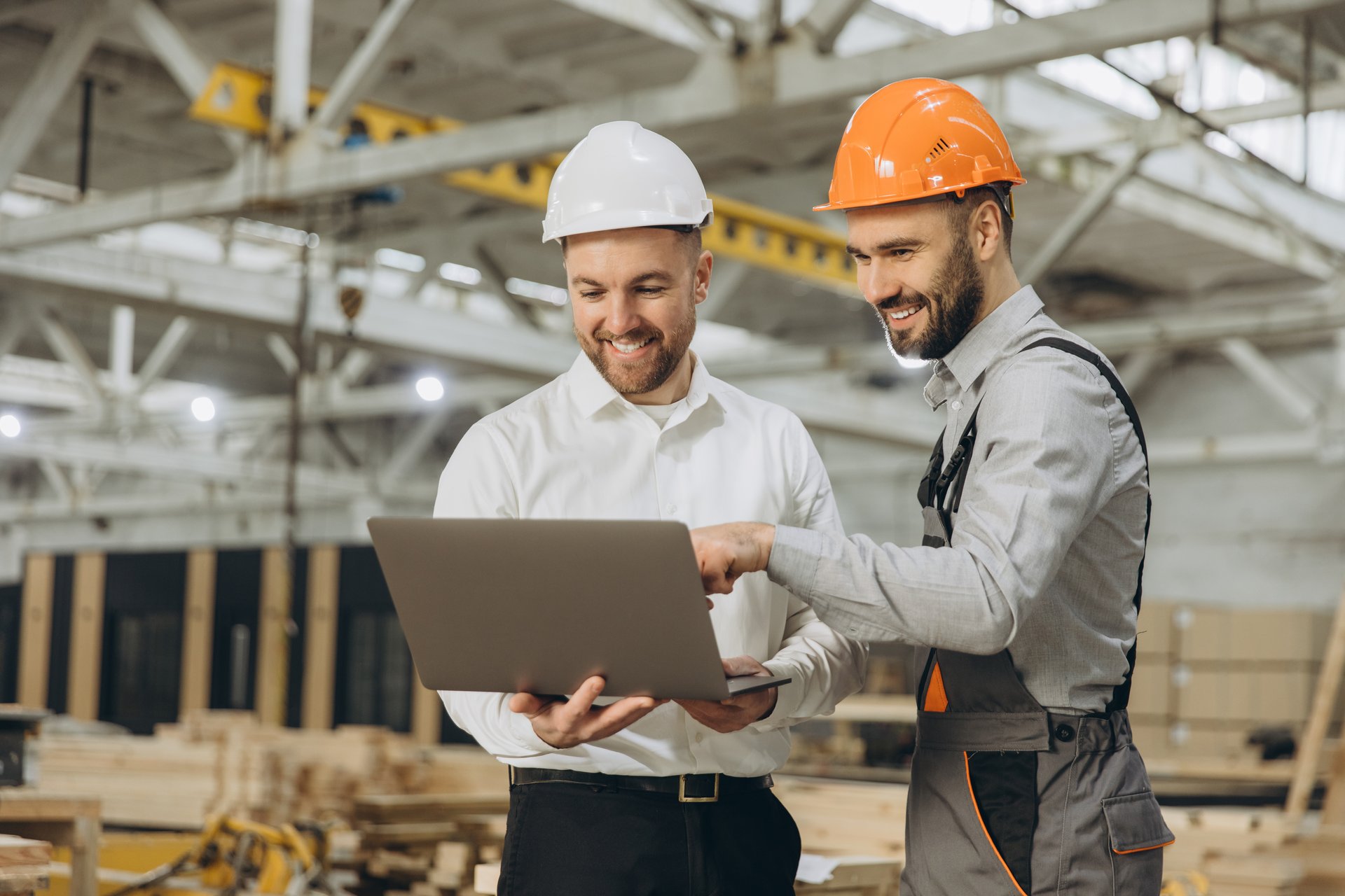 Two engineers collaborate on a laptop in a busy factory, discussing a project. They focus on sustainable, eco-friendly construction, using modern tech for efficiency