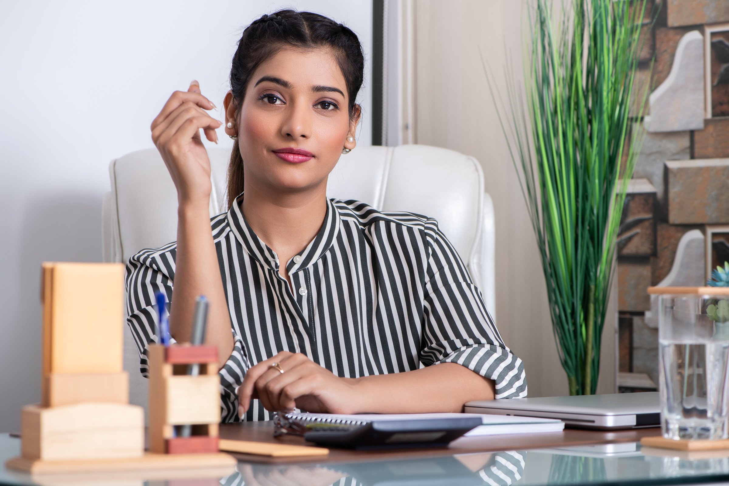 A poised and charismatic Chief Executive Officer of a leading consultancy firm, she exudes confidence and elegance. Light makeup and a touch of red lipstick enhance her natural charm, while her hair is neatly clipped back, completing her polished, professional look. Dressed in a crisp white and black striped formal shirt, she sits gracefully on a sleek white chair within her office cabin.