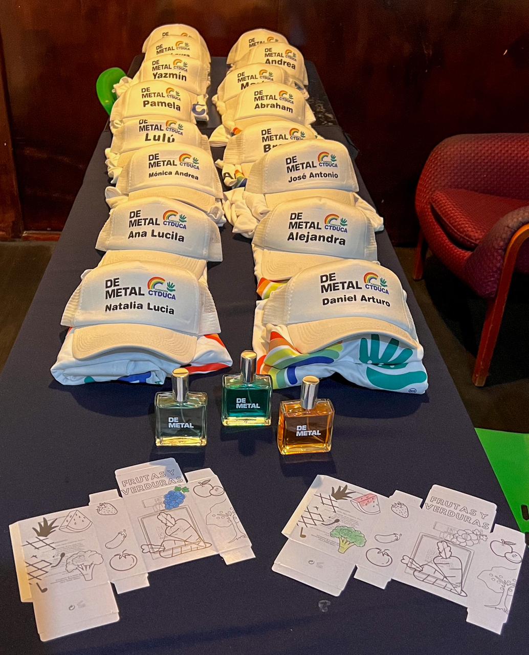 Rows of white caps with names and colorful bottles on a table. Caps have "DE METAL GUACIA" text and fruit drawings underneath.