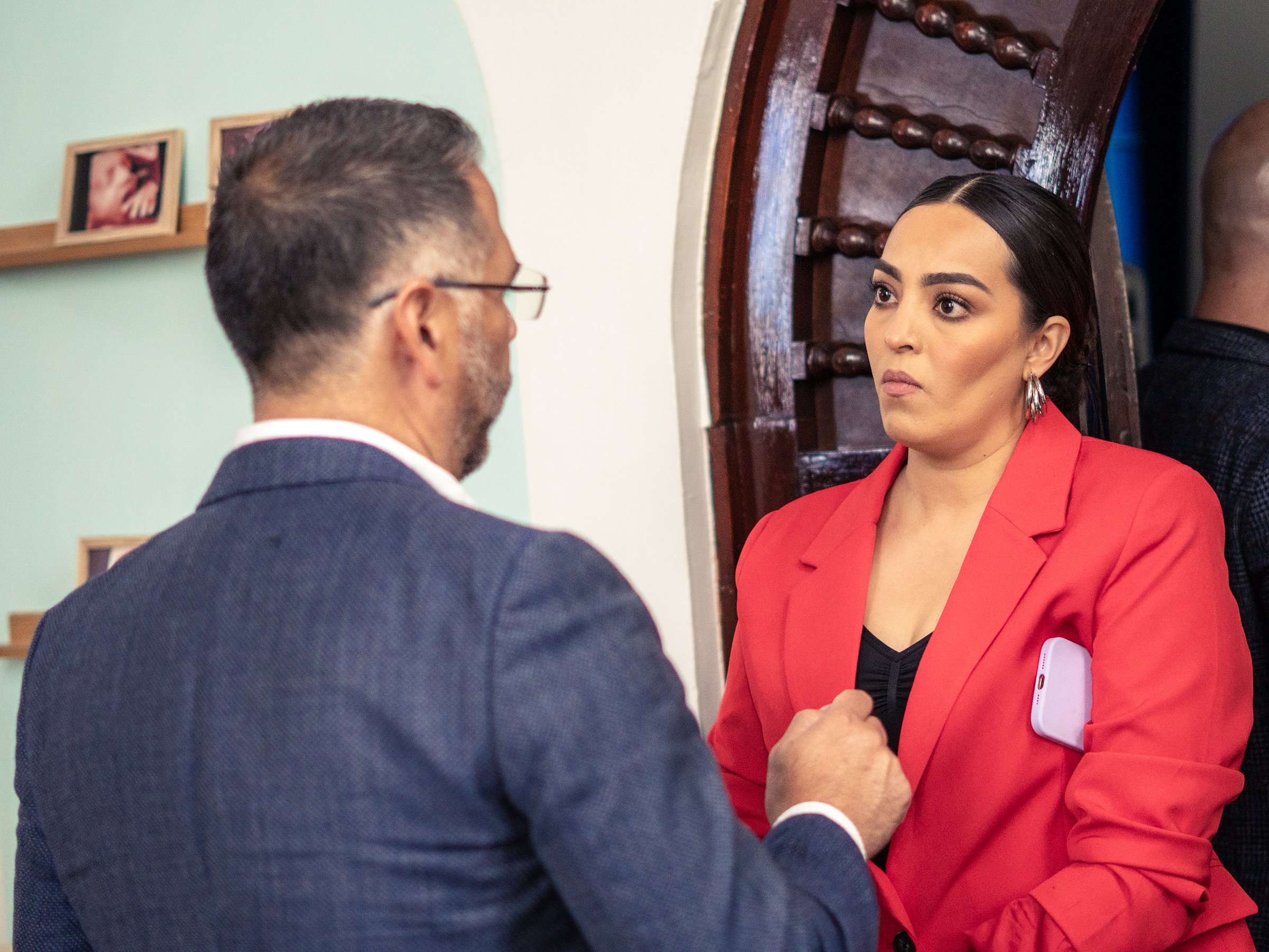 Two people having a serious conversation indoors, one wearing a red blazer and the other in a suit, standing near a wooden door.