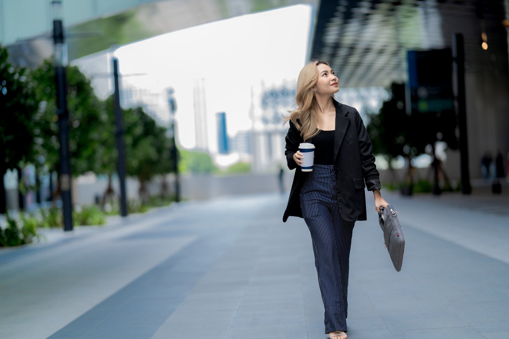 Businesswoman standing in a modern urban setting, holding her bag with a confident expression, representing professionalism, elegance, and readiness in a sleek and dynamic environment.