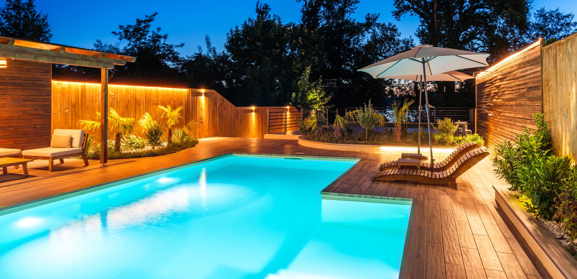 A beautifully lit pool with comfortable loungers, umbrellas, and lush greenery surrounding the deck, creating an inviting atmosphere at night. Concept of luxury outdoor relaxation and serene nighttime ambiance