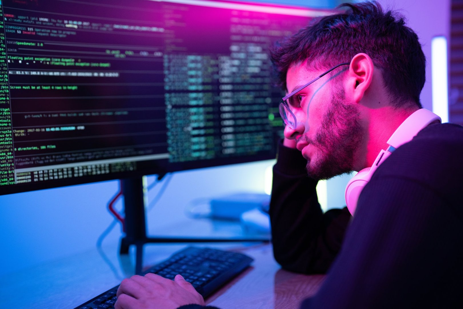 In a low-light environment, an individual focuses intensely on coding, surrounded by dual-screen monitors filled with complex computer code. The colorful ambient lighting enhances the atmosphere.