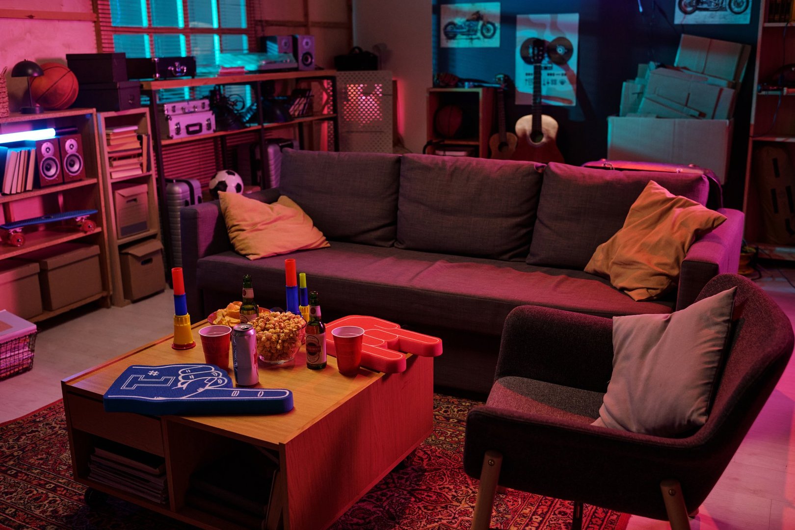 Living room scene showing empty sofa and armchair with table holding snacks, beer bottles, party cups, foam finger, party horns, illuminated by colorful lights, sports equipment in background