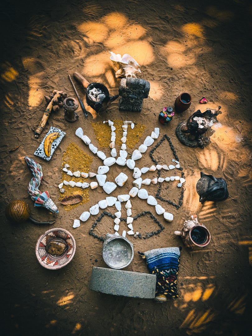 An artistic arrangement of stones, figurines, and pottery on sandy ground, forming a circular pattern with a central star shape.