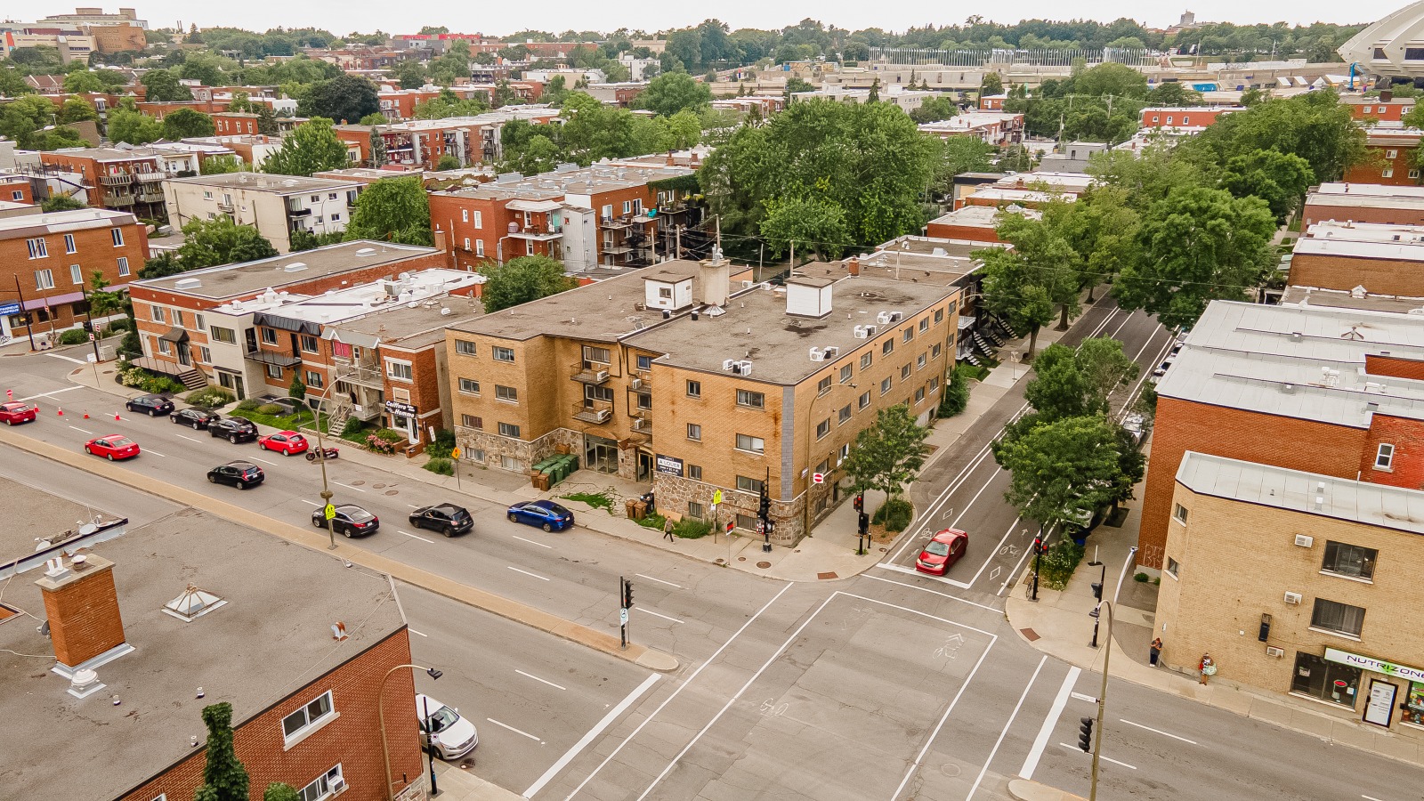 Aerial view of an urban neighborhood with brick buildings, streets, and parked cars, surrounded by greenery and trees.