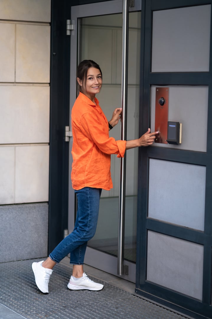 Brunette woman wearing orange shirt and jeans dialing an intercom to enter a building.