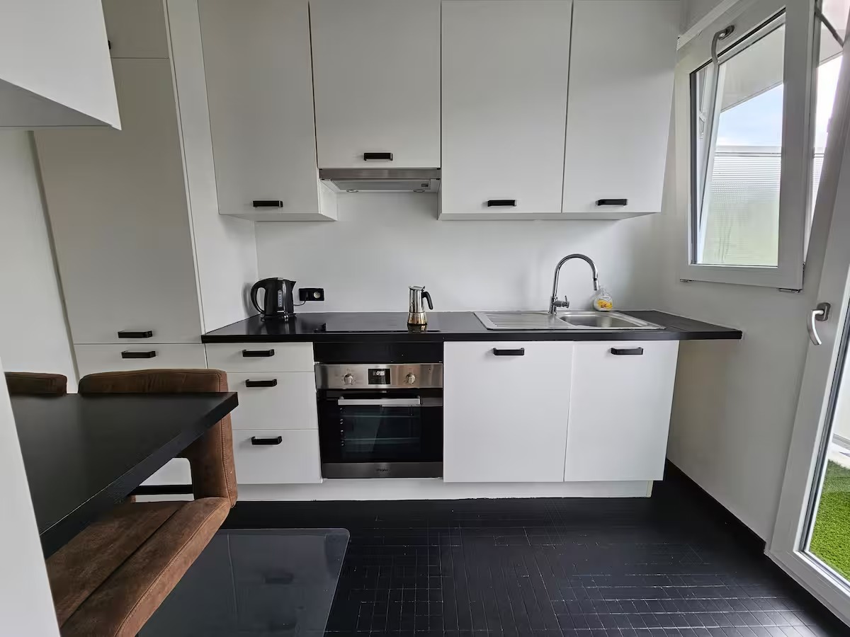 Modern white kitchen with black countertops, stove, stainless steel sink, and a window. Includes a kettle and espresso maker.