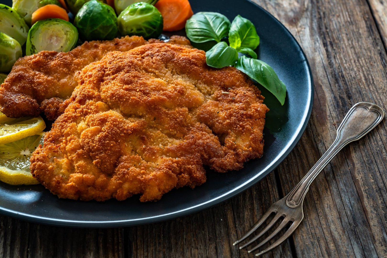 Crispy breaded fried pork chop, boiled potatoes and fresh vegetables served on wooden table