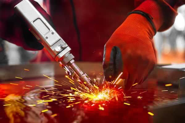 A strong man in a welders leathers, a metal product is welded with a welding machine in the garage, fire sparkles fly to the sides.