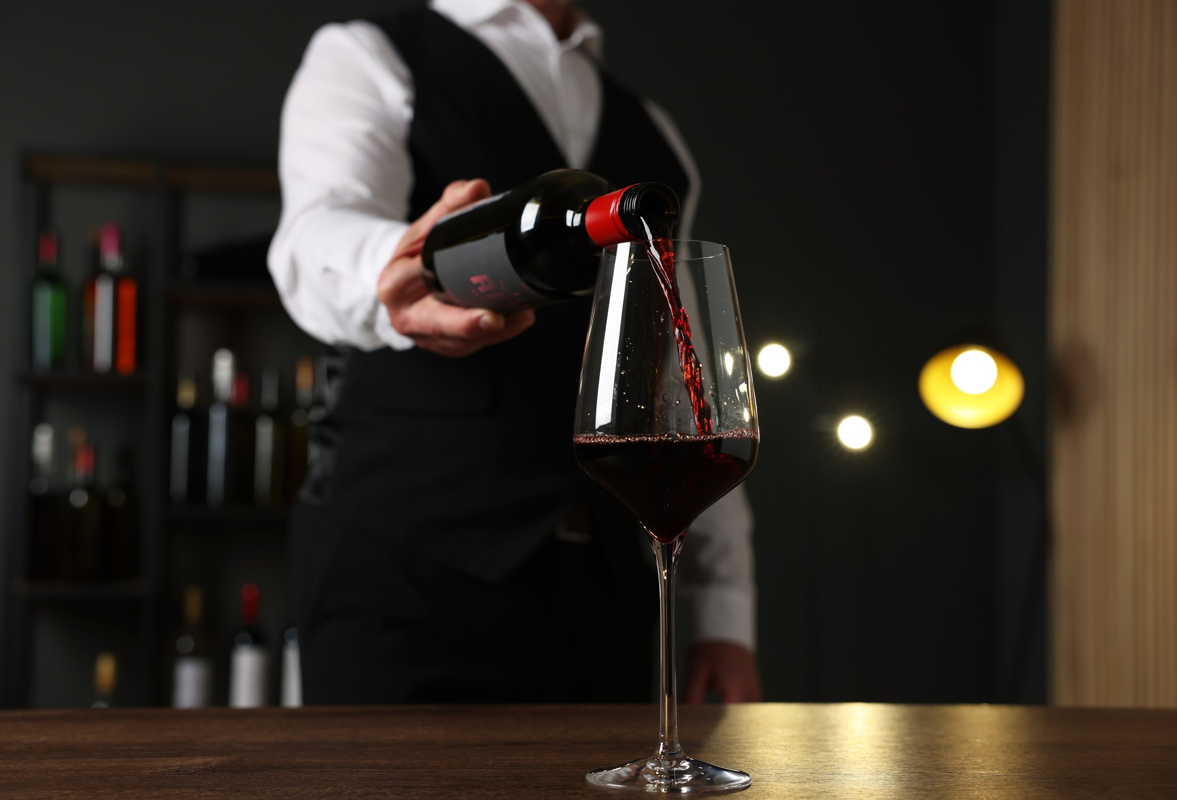 Professional sommelier pouring red wine into glass at wooden table indoors, closeup