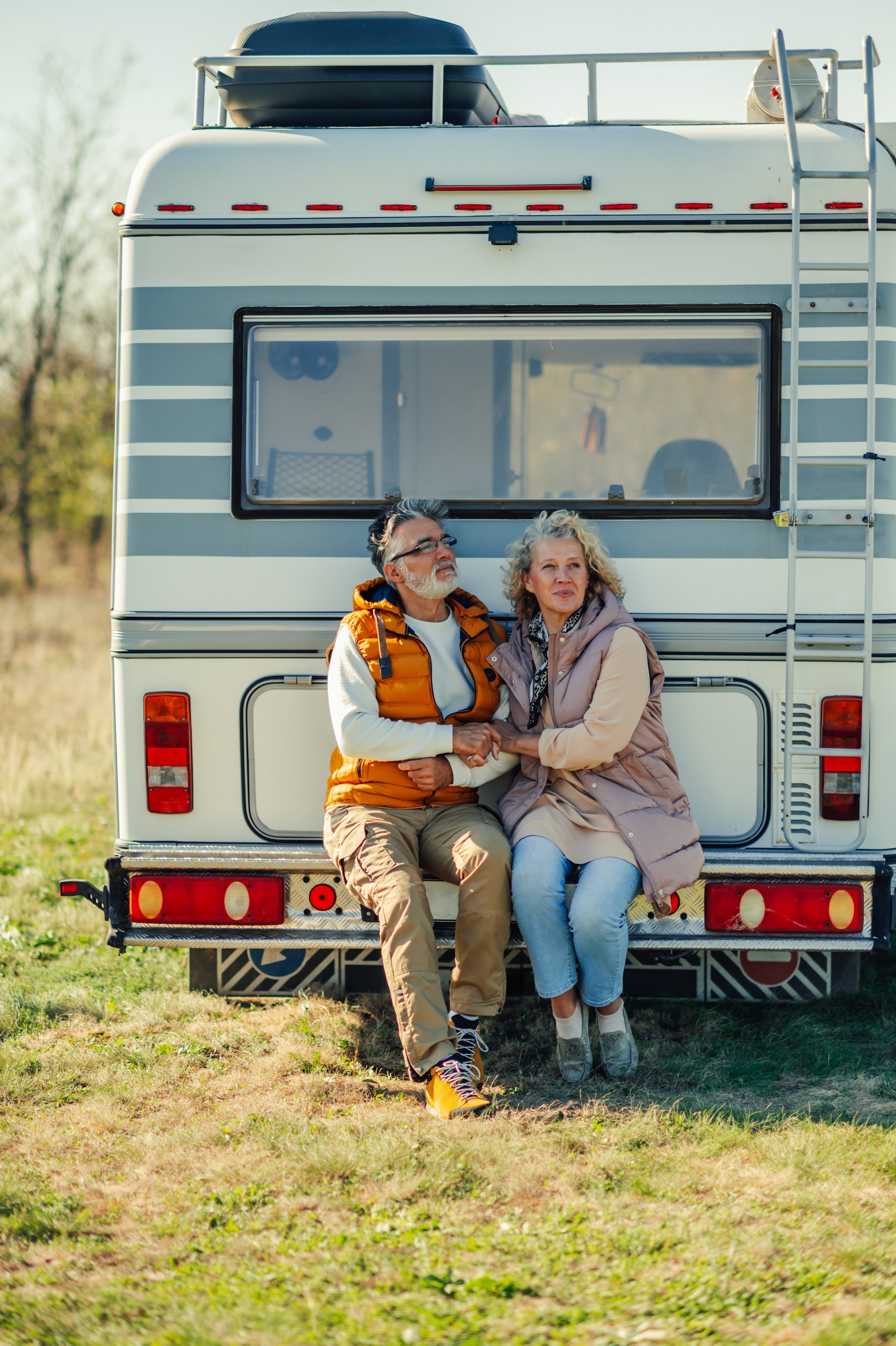 Happy retired couple sitting on back of their camper van, holding hands and enjoying freedom of travel and adventure in nature